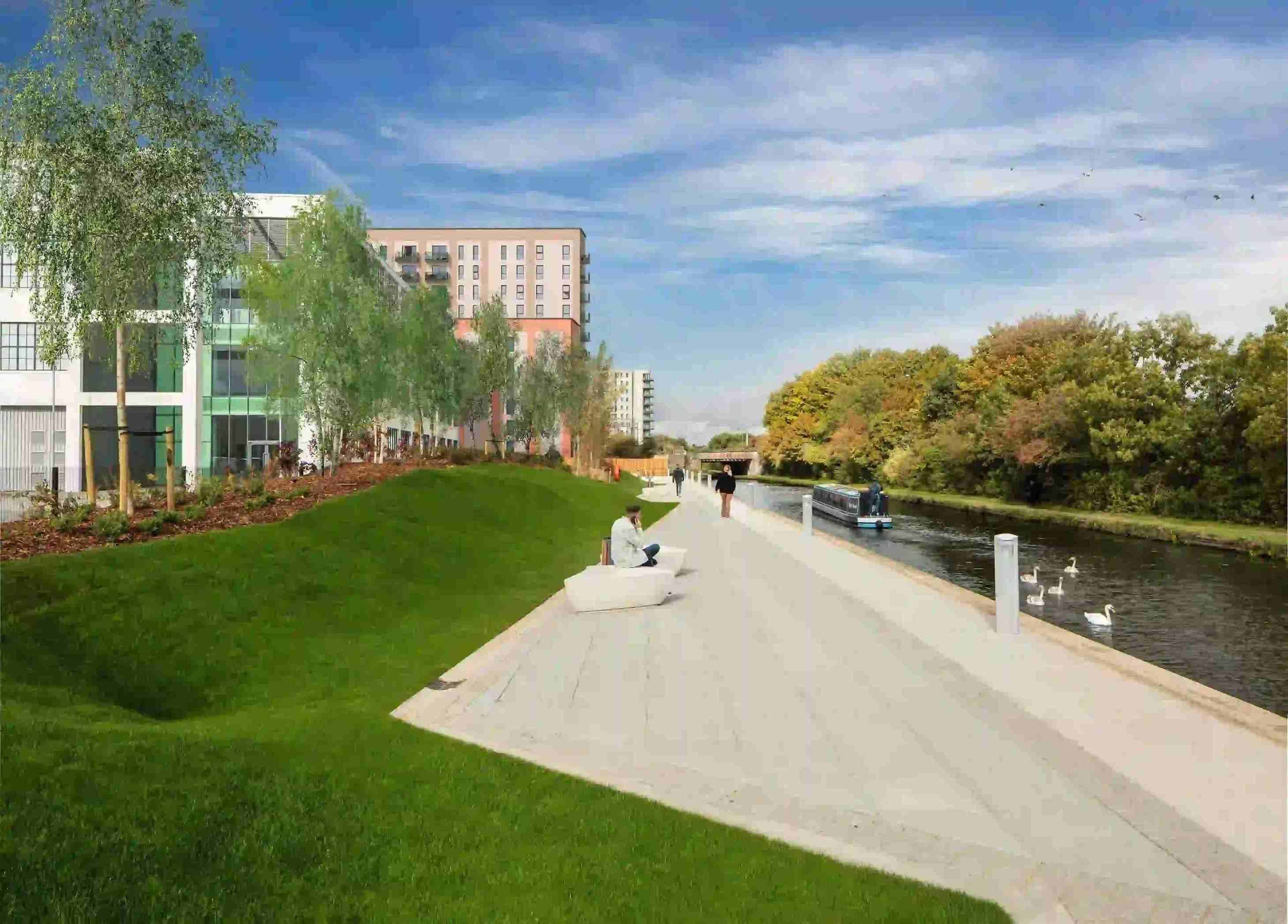 A grassy path next to a Hayes Village canal with buildings in the background.